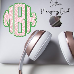 May include: A custom monogram decal with a green outline and pink zebra print letters. The decal is on a white surface next to a pair of white and rose gold headphones. The text "Custom Monogram Decal" is written in cursive.