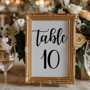May include: A gold framed table number sign with the word "Table" in elegant script above the number "10" in a bold font. The sign is displayed on a table with floral arrangements and glassware in the background.