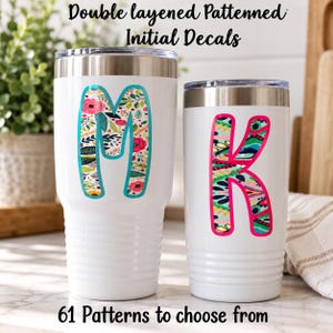 May include: Two white stainless steel tumblers with metal lids. The larger tumbler features a floral patterned letter M with a teal outline. The smaller tumbler features a colorful abstract patterned letter K with a hot pink outline.