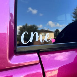 May include: White vinyl decal with the word "CMe" in a cursive font, accompanied by a small pink heart. The decal is affixed to a car window, with a bright pink car body visible. The background shows a blue sky and trees.