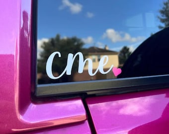May include: A white vinyl decal applied to a vehicle window featuring the lowercase cursive text cme followed by a small solid pink heart. The decal is positioned on the glass of a bright magenta car door, visible against a blue sky background.