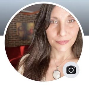 May include: A woman with long, dark hair and a silver pendant necklace. The pendant is oval-shaped with a light-colored stone. The background includes a brick wall, a painting, and a red couch. A camera icon is in the lower right corner.