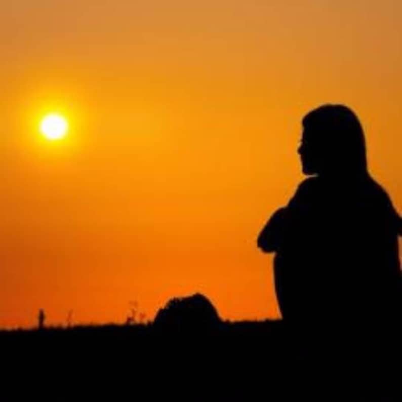 May include: Silhouette of a person sitting with their arms crossed, looking out at a bright orange sunset.