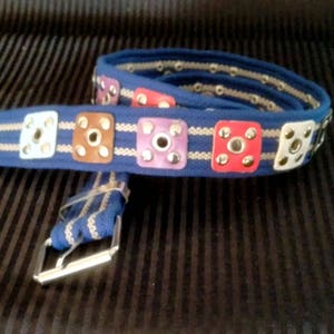 Peut inclure: Une ceinture en tissu bleu avec des accents carrés décoratifs de différentes couleurs, dont blanc, marron, rouge et violet. La ceinture a une boucle argentée et des œillets sur les bords. Un accessoire élégant.