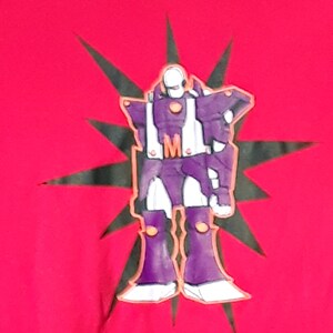 Pu&ograve; includere: T-shirt rossa con un disegno di robot viola, dettagli bianchi e la lettera "M". Il robot &egrave; posto su un motivo a stella nera.