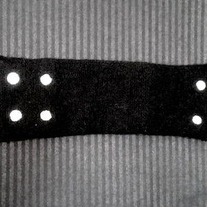 May include: A black rectangular headband with five white reflective dots. The headband is made of a soft, textured material and is designed to be worn on the head. The background is a gray striped fabric.