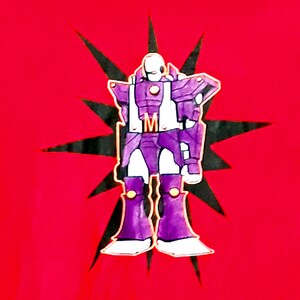 Pu&ograve; includere: T-shirt rossa con un robot dei cartoni animati viola, bianco e oro. Il robot &egrave; posto su una stella nera. Il robot ha la lettera "M" sul petto. Una maglietta grafica.