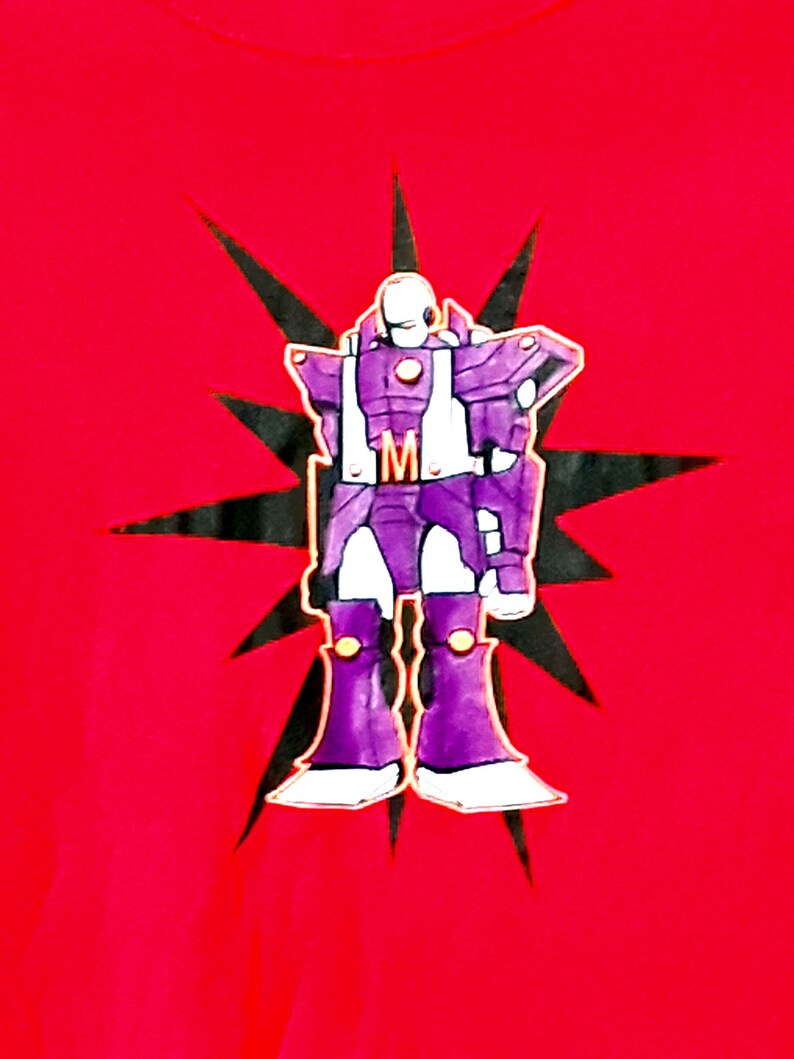 Pu&ograve; includere: T-shirt rossa con un robot dei cartoni animati viola, bianco e oro. Il robot &egrave; posto su una stella nera. Il robot ha la lettera "M" sul petto. Una maglietta grafica.