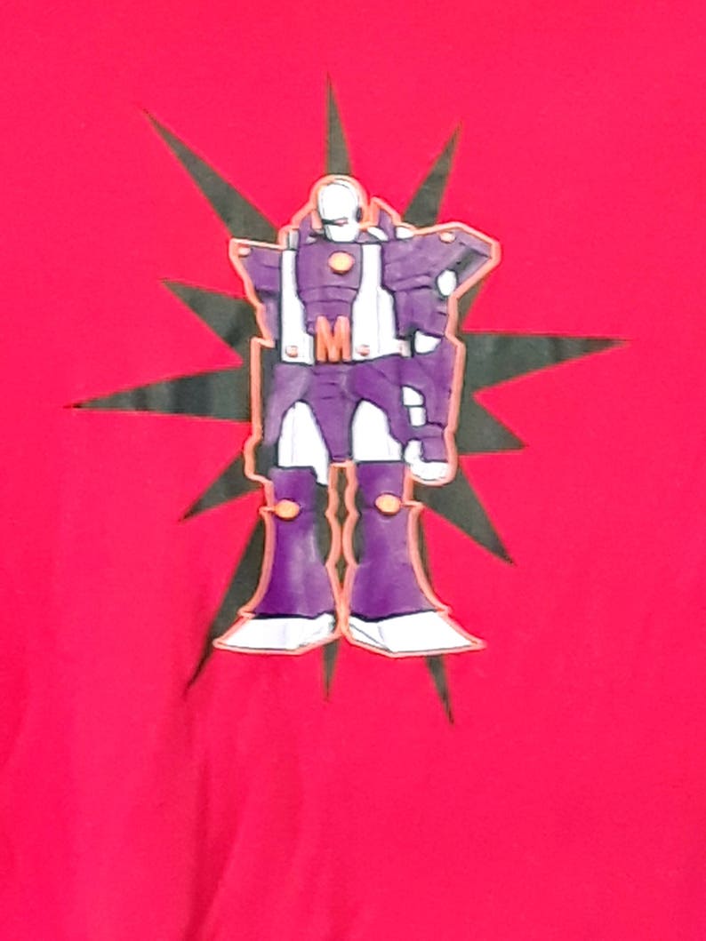 Pu&ograve; includere: Maglietta rossa con un disegno di robot viola, dettagli bianchi e arancioni e una stella nera. Il robot ha la lettera "M" sul petto. Il disegno &egrave; centrato.