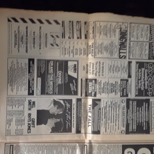 May include: A vintage newspaper page featuring numerous black and white advertisements. The ads promote live music events, including bands like Divine, Dr. and the Medics, and The Fall. The page is aged with visible wear and tear.