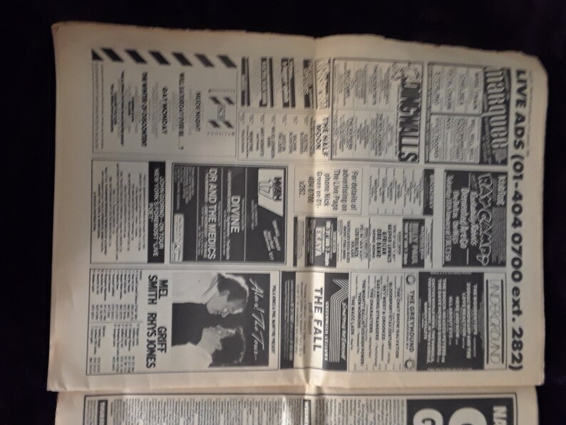 May include: A vintage newspaper page featuring numerous black and white advertisements. The ads promote live music events, including bands like Divine, Dr. and the Medics, and The Fall. The page is aged with visible wear and tear.