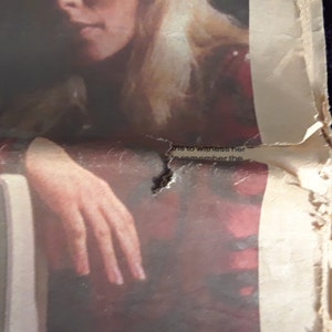 May include: A torn, aged newspaper page with a partial image of a person and a hand resting on a surface. The paper is off-white with red and black ink, and the edges are tattered. Faint text is visible near the top.