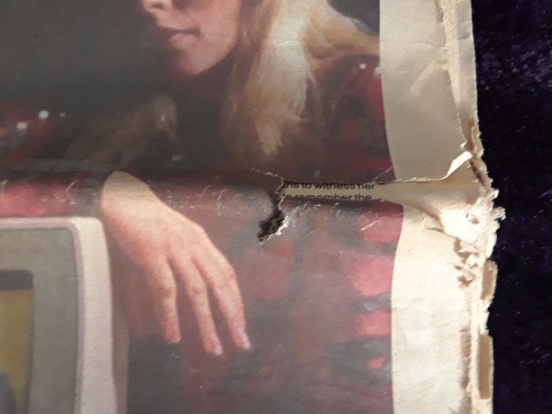 May include: A torn, aged newspaper page with a partial image of a person and a hand resting on a surface. The paper is off-white with red and black ink, and the edges are tattered. Faint text is visible near the top.
