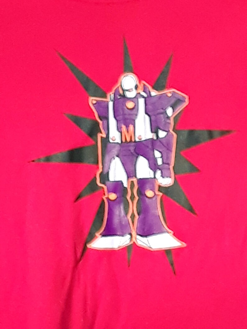 Pu&ograve; includere: T-shirt rossa con un disegno di robot viola, dettagli bianchi e la lettera "M". Il robot &egrave; posto su un motivo a stella nera.