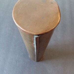 Puede incluir: Un recipiente cil&iacute;ndrico de color cobre con una parte superior redondeada. El recipiente se estrecha ligeramente hacia la parte inferior. El metal tiene un tono c&aacute;lido, marr&oacute;n rojizo y muestra signos de edad y uso. La parte superior es un c&iacute;rculo plano y s&oacute;lido.