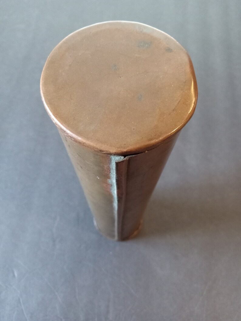 Puede incluir: Un recipiente cil&iacute;ndrico de color cobre con una parte superior redondeada. El recipiente se estrecha ligeramente hacia la parte inferior. El metal tiene un tono c&aacute;lido, marr&oacute;n rojizo y muestra signos de edad y uso. La parte superior es un c&iacute;rculo plano y s&oacute;lido.
