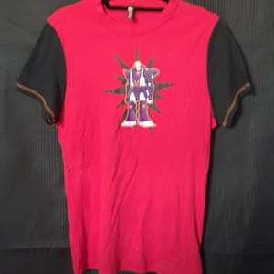 Pu&ograve; includere: T-shirt rosso con maniche nere e una grafica di una figura simile a un robot. Il robot &egrave; viola, bianco e oro, su un design a stella verde. Le maniche hanno cuciture arancioni. La maglietta &egrave; appesa a una gruccia nera.