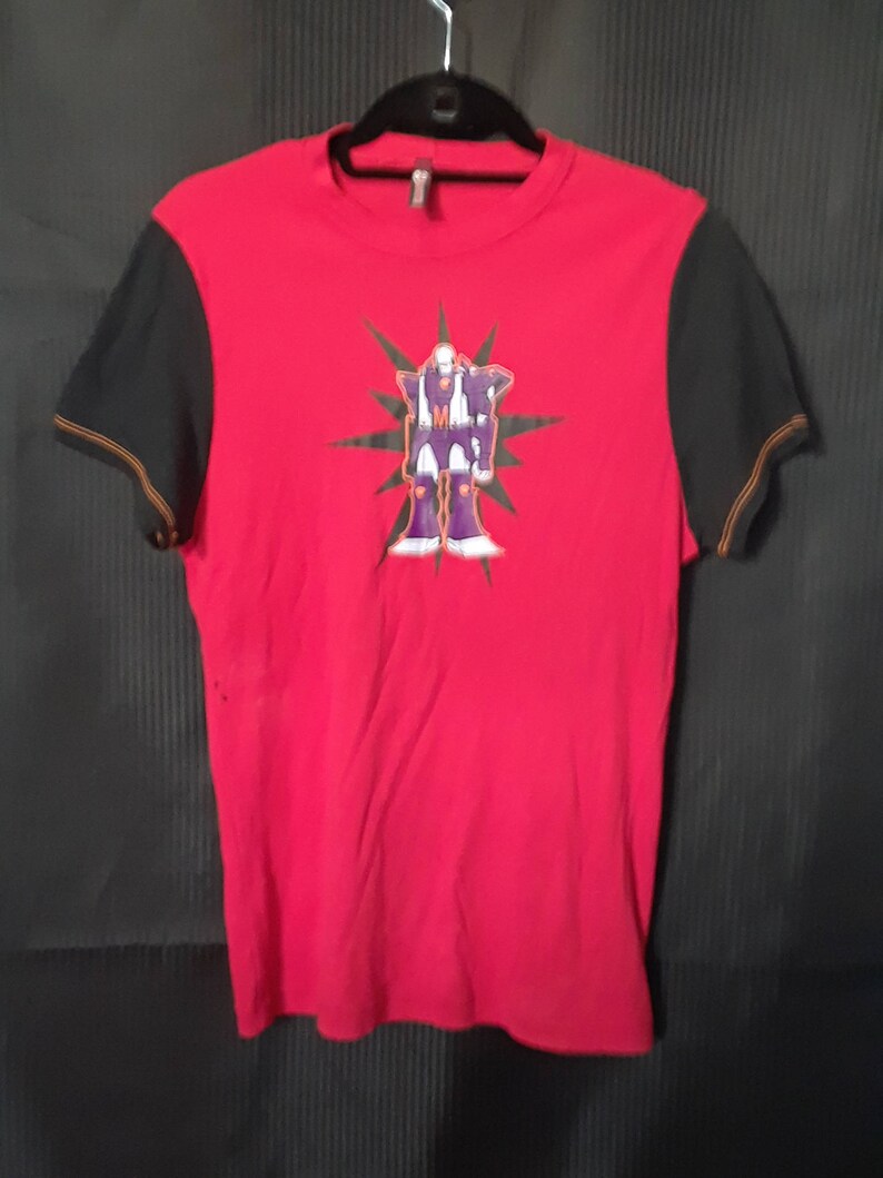 Pu&ograve; includere: T-shirt rosso con maniche nere e una grafica di una figura simile a un robot. Il robot &egrave; viola, bianco e oro, su un design a stella verde. Le maniche hanno cuciture arancioni. La maglietta &egrave; appesa a una gruccia nera.