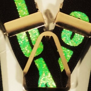 May include: Black suspenders with gold-coloured metal clips and a central adjuster. The suspenders feature the word "LOVE" in bright green glitter. The background is a neutral white.