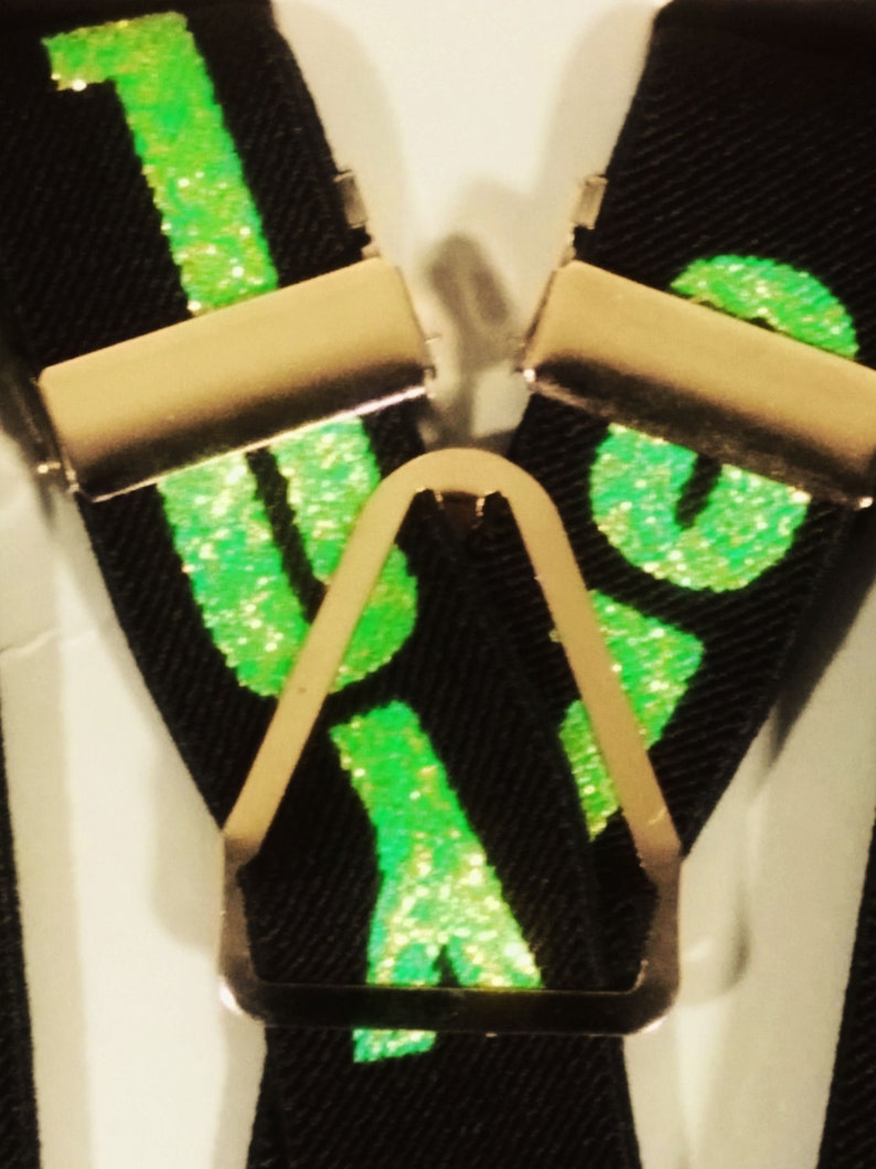 May include: Black suspenders with gold-coloured metal clips and a central adjuster. The suspenders feature the word "LOVE" in bright green glitter. The background is a neutral white.