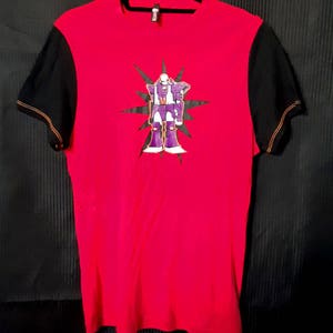 Pu&ograve; includere: Maglietta rossa con maniche nere e cuciture arancioni. La parte anteriore presenta una grafica di robot viola e bianca con un motivo a stella. La maglietta &egrave; appesa a una gruccia nera su sfondo nero.