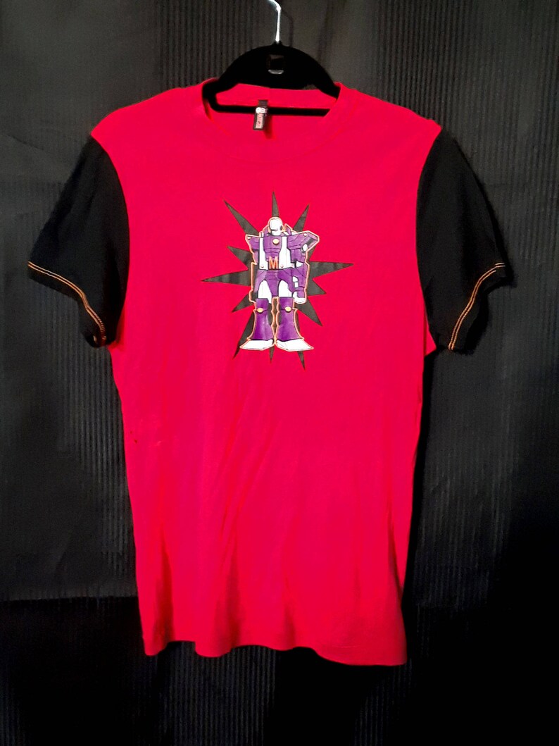 Pu&ograve; includere: Maglietta rossa con maniche nere e cuciture arancioni. La parte anteriore presenta una grafica di robot viola e bianca con un motivo a stella. La maglietta &egrave; appesa a una gruccia nera su sfondo nero.