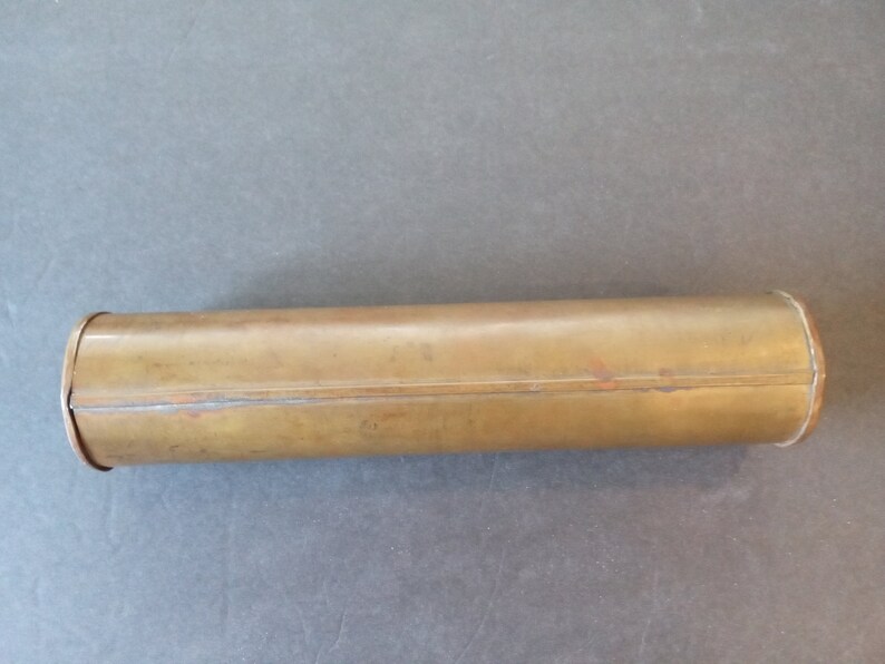 Puede incluir: Un recipiente cil&iacute;ndrico de color lat&oacute;n con extremos redondeados. El recipiente tiene una costura a lo largo y muestra signos de desgaste. La forma es larga y estrecha, de unos 20 cm de largo, posiblemente un tubo de almacenamiento.