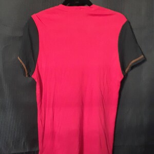 Pu&ograve; includere: Una maglietta rossa con maniche nere appesa a una gruccia nera. Le maniche hanno cuciture arancioni lungo i bordi. La maglietta &egrave; realizzata in un materiale morbido ed &egrave; probabilmente per abbigliamento casual.