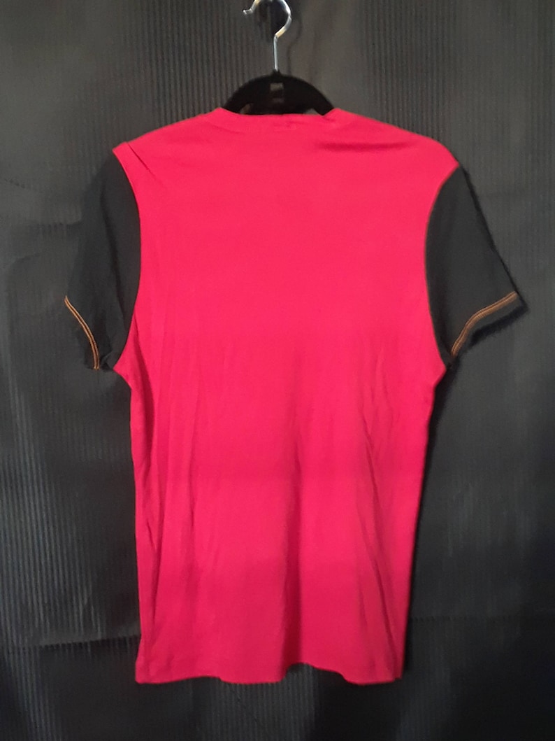 Pu&ograve; includere: Una maglietta rossa con maniche nere appesa a una gruccia nera. Le maniche hanno cuciture arancioni lungo i bordi. La maglietta &egrave; realizzata in un materiale morbido ed &egrave; probabilmente per abbigliamento casual.