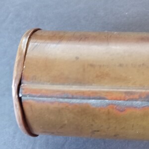 Puede incluir: Primer plano de un recipiente cil&iacute;ndrico de color cobre. El recipiente tiene un borde redondeado y una costura que recorre su longitud. El metal tiene un aspecto ligeramente envejecido con variaciones de color.