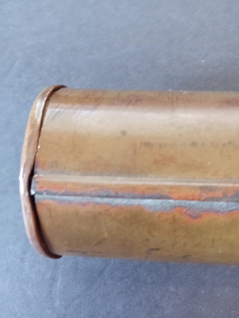 Puede incluir: Primer plano de un recipiente cil&iacute;ndrico de color cobre. El recipiente tiene un borde redondeado y una costura que recorre su longitud. El metal tiene un aspecto ligeramente envejecido con variaciones de color.