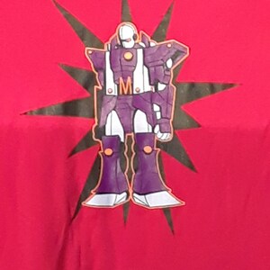 Pu&ograve; includere: T-shirt rossa con un design di robot viola, dettagli bianchi e arancioni e una stella nera. Il robot ha la lettera "M" sul petto. Una maglietta grafica.