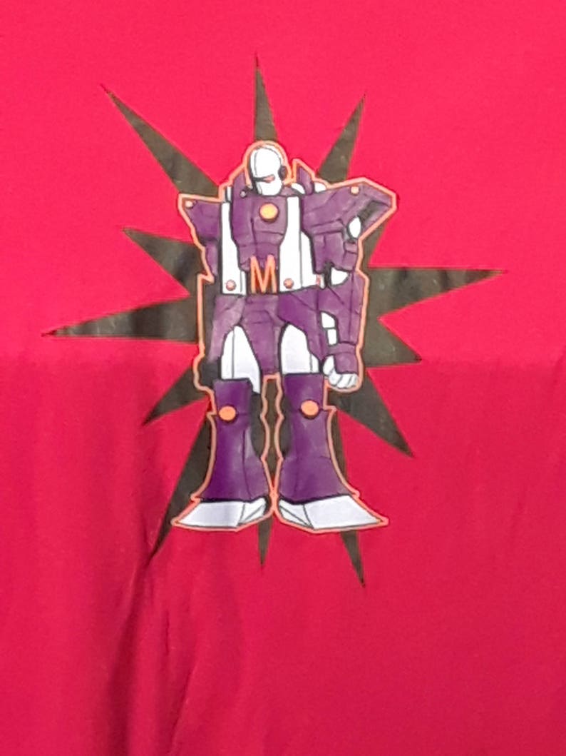 Pu&ograve; includere: T-shirt rossa con un design di robot viola, dettagli bianchi e arancioni e una stella nera. Il robot ha la lettera "M" sul petto. Una maglietta grafica.