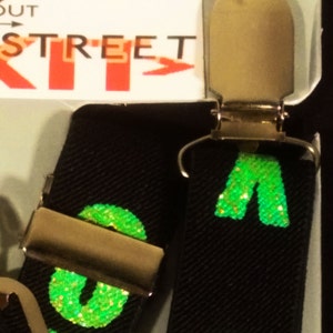 May include: Black suspenders with gold-coloured metal clips. The suspenders feature a repeating pattern of green glitter letters. The word "STREET" is visible on the packaging.