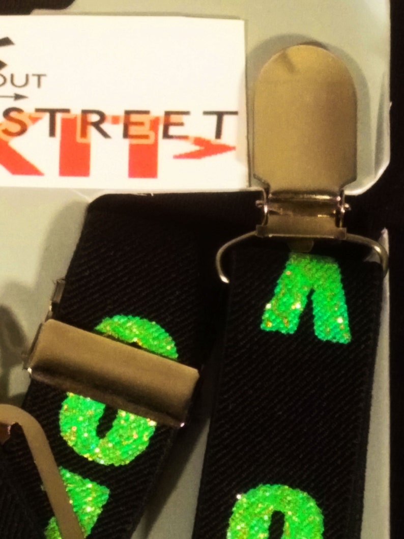 May include: Black suspenders with gold-coloured metal clips. The suspenders feature a repeating pattern of green glitter letters. The word "STREET" is visible on the packaging.