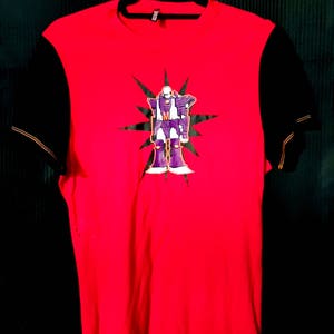 Pu&ograve; includere: T-shirt rosso con maniche nere. La parte anteriore presenta una grafica di un robot viola con dettagli bianchi e un motivo a stella. La maglietta &egrave; appesa a una gruccia nera.