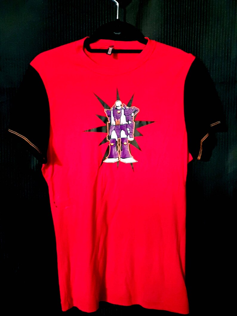 Pu&ograve; includere: T-shirt rosso con maniche nere. La parte anteriore presenta una grafica di un robot viola con dettagli bianchi e un motivo a stella. La maglietta &egrave; appesa a una gruccia nera.
