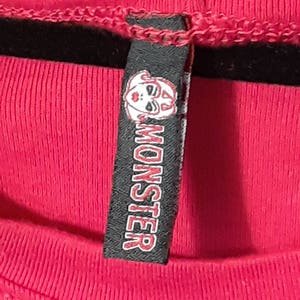 Pu&ograve; includere: Primo piano di una maglietta rosa acceso con un'etichetta nera. L'etichetta presenta una grafica di un volto bianco e la parola "MONSTER" in lettere rosse. La maglietta ha una trama a coste.