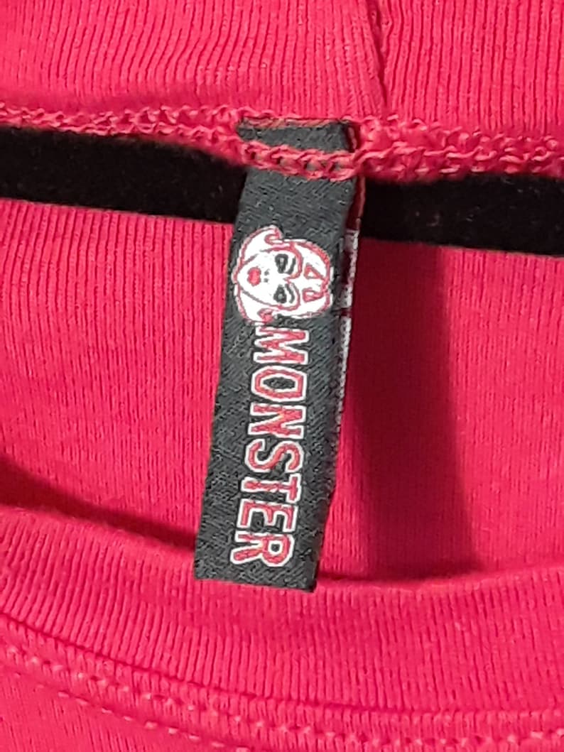Pu&ograve; includere: Primo piano di una maglietta rosa acceso con un'etichetta nera. L'etichetta presenta una grafica di un volto bianco e la parola "MONSTER" in lettere rosse. La maglietta ha una trama a coste.
