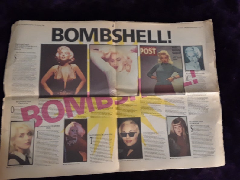 May include: A newspaper spread with the headline "BOMBSHELL!" in large black letters. The pages feature images of blonde women, including Marilyn Monroe, and the word "BOMB" in pink. The newspaper is on a dark purple surface.