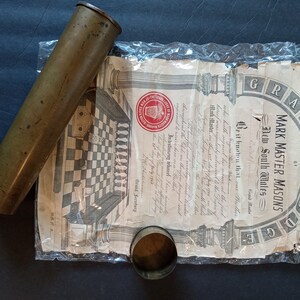 Puede incluir: Un cilindro de lat&oacute;n antiguo y una peque&ntilde;a taza de metal descansan junto a un documento. El documento, parcialmente visible, presenta las palabras "MARK MASTER MASONS" y "New South Wales" con una ilustraci&oacute;n.