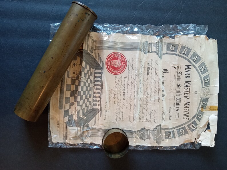Puede incluir: Un cilindro de lat&oacute;n antiguo y una peque&ntilde;a taza de metal descansan junto a un documento. El documento, parcialmente visible, presenta las palabras "MARK MASTER MASONS" y "New South Wales" con una ilustraci&oacute;n.