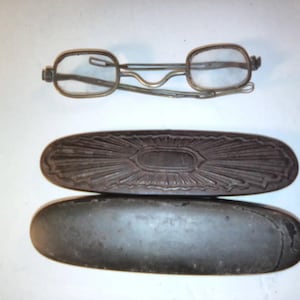 May include: A pair of vintage metal spectacles with thin frames and clear lenses, positioned above an oval-shaped dark brown case featuring a decorative sunburst pattern on the lid and a smooth, solid base.
