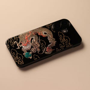 以下が含まれることがあります： 黒い携帯電話ケース。金色の龍のデザイン、赤いアクセント、ターコイズの宝石が特徴です。 龍は複雑に詳細で、ケースは光沢のある仕上がりです。 デザインは金色の雲の背景に設定されています。