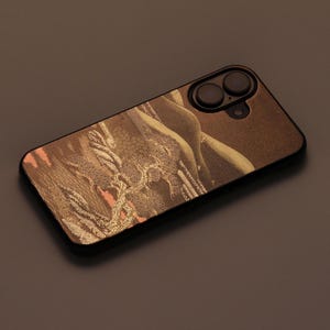 Può includere: Una custodia per telefono nera con un design dorato e marrone che presenta un paesaggio in stile giapponese con un albero e montagne. La custodia ha un ritaglio per l'obiettivo della fotocamera.