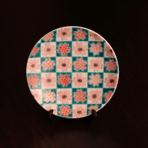 May include: A round ceramic plate featuring a checkerboard pattern of alternating teal squares and white squares. The white squares are decorated with pink cherry blossom flowers, while the teal squares contain red geometric patterns.