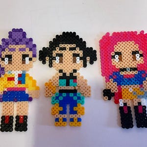 Può includere: Tre figure colorate in pixel art. Le figure sono realizzate con piccole perline fuse. Una figura ha i capelli viola, un top giallo e una gonna blu. Un'altra ha i capelli neri, un top turchese e pantaloni blu. La terza ha i capelli rosa, un top blu e una gonna gialla.