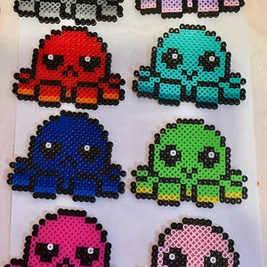 May include: A collection of eight colorful octopus figures made from fused plastic beads. Each octopus has a round head, eight tentacles, and two white eyes. The octopuses are in various colors, including gray, purple, red, teal, blue, green, pink, and light pink.