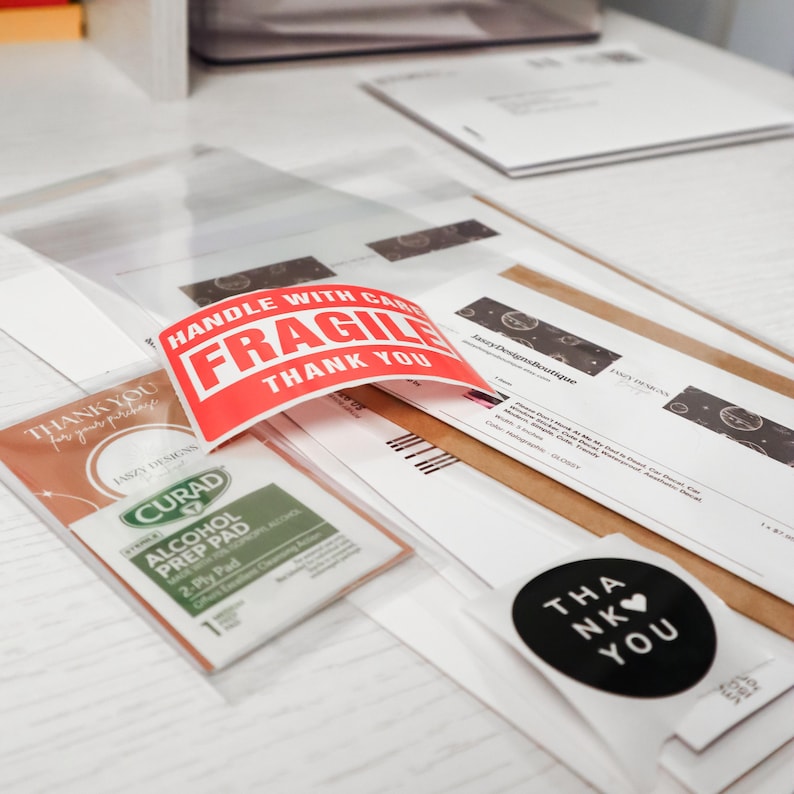 May include: A collection of shipping materials including a red and white sticker with the text HANDLE WITH CARE FRAGILE THANK YOU, a Curad alcohol prep pad, a circular black sticker with the text THANK YOU, and various printed order documents.