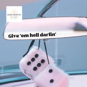 May include: A rearview mirror with the text Give 'em hell darlin' printed in black on the surface. Hanging from the mirror are two fuzzy light pink dice with black circular pips, suspended by thin black cords against a light blue background.